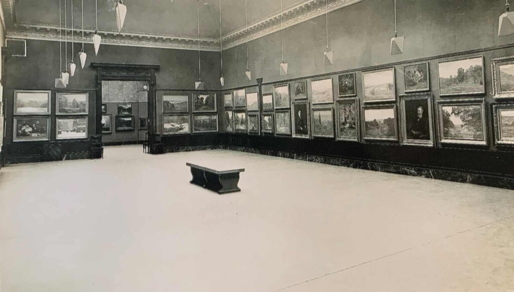 many paintings line the wall of an otherwise empty gallery room with a single bench in the center