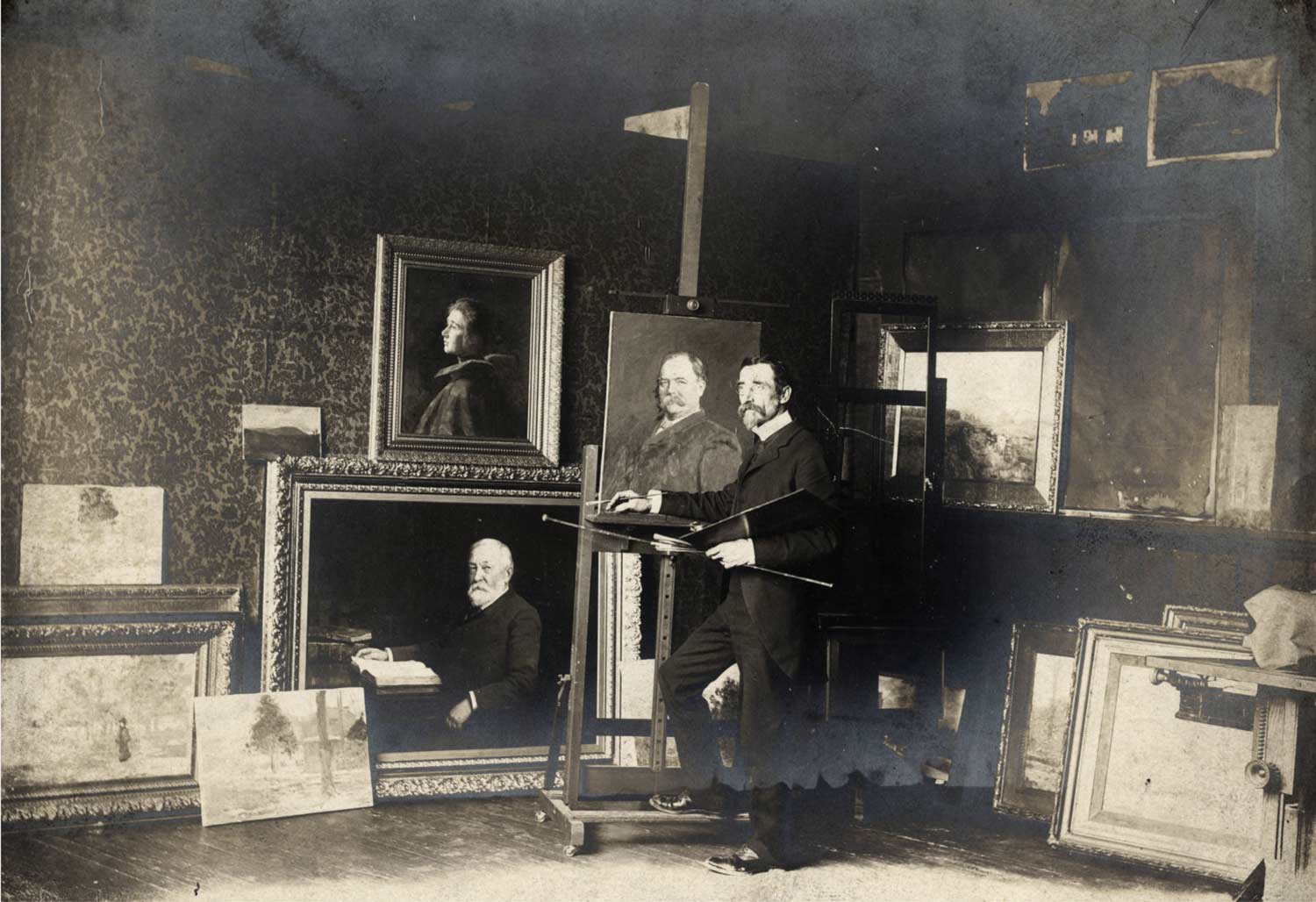 The artist stands with palette and brush in hand in front of a portrait on an easel. many paintings surround him.