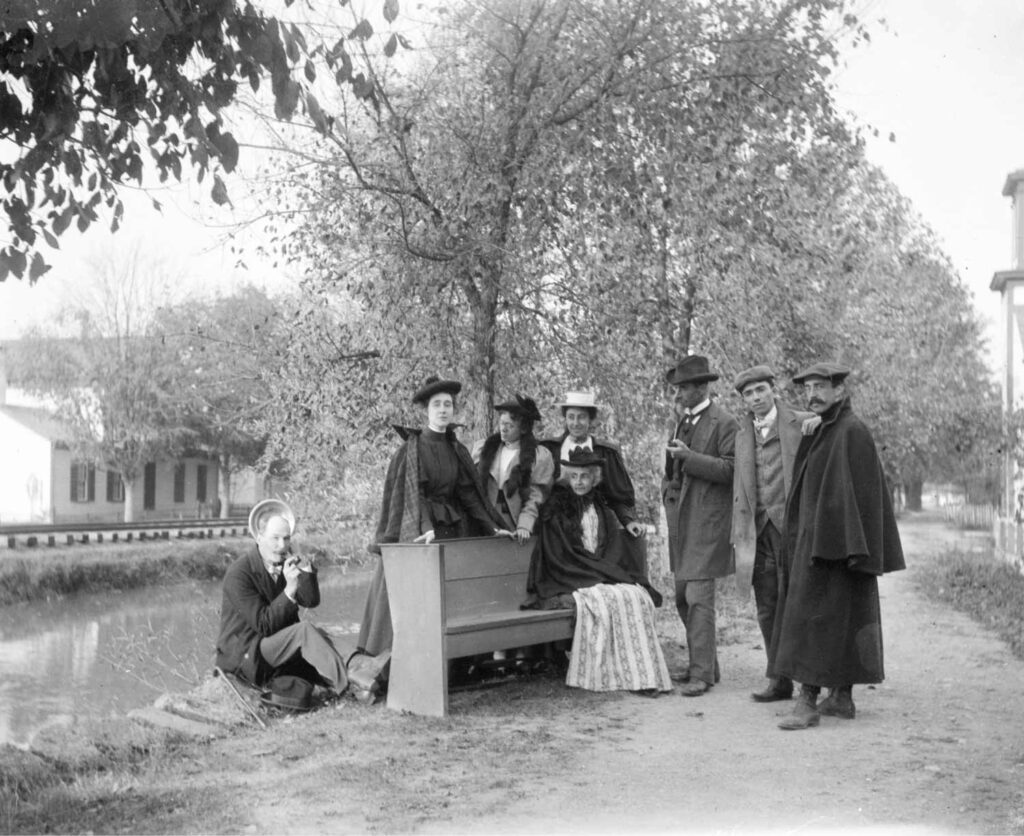 black and white photograph of 8 men and women in fine clothes, hats, and capes or jackets posing in front of a canal.